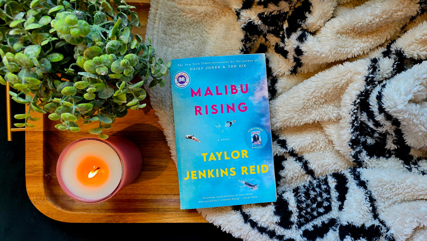 Book titled 'Malibu Rising' by Taylor Jenkins Reid on a wooden tray with a candle and plant, on a textured surface.
