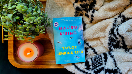 Book titled 'Malibu Rising' by Taylor Jenkins Reid on a wooden tray with a candle and plant, on a textured surface.