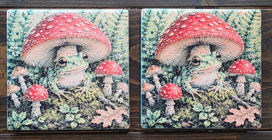 Two square ceramic coasters with a whimsical illustration of a woman's face hidden under a large red-and-white mushroom, surrounded by green foliage and small red mushrooms, displayed on a dark wood background.